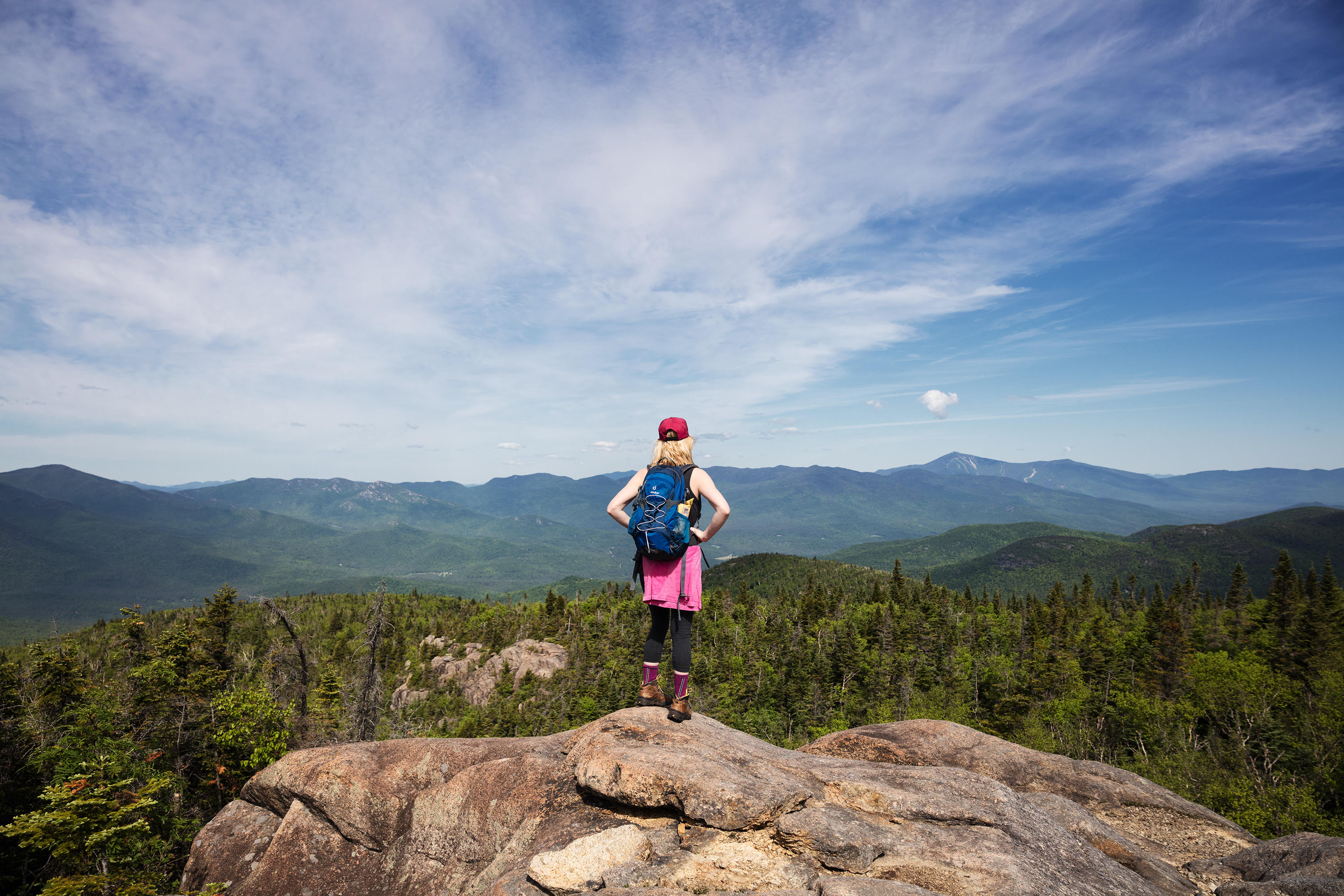 A woman in hiking gear facing away from camera looks out over mountains, forest and sky.